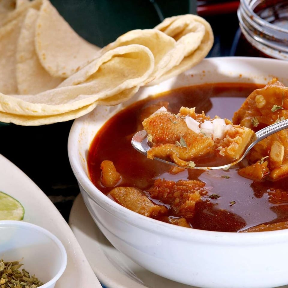 Bowl of meat in red chili sauce with tortillas, lime wedges, and a spoonful of meat garnished with onion.
