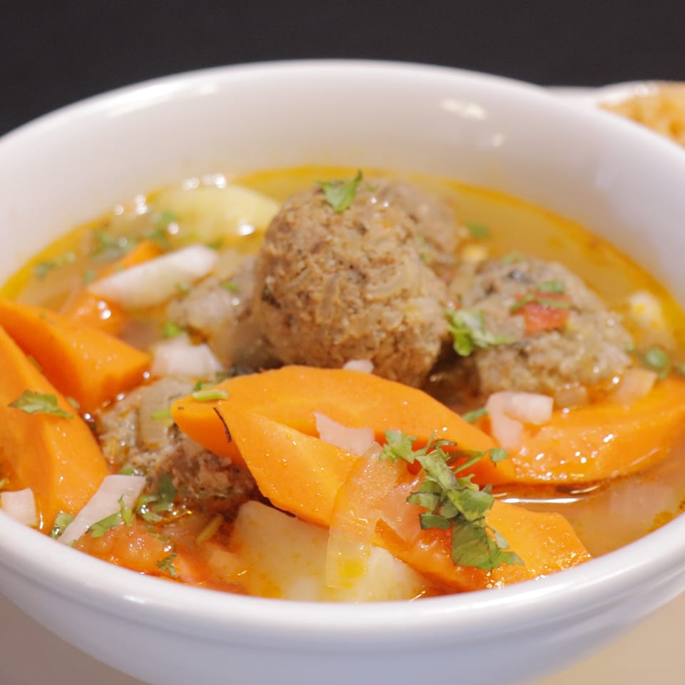 Bowl of meatballs and vegetables in golden broth, garnished with cilantro, served alongside a dish of rice.