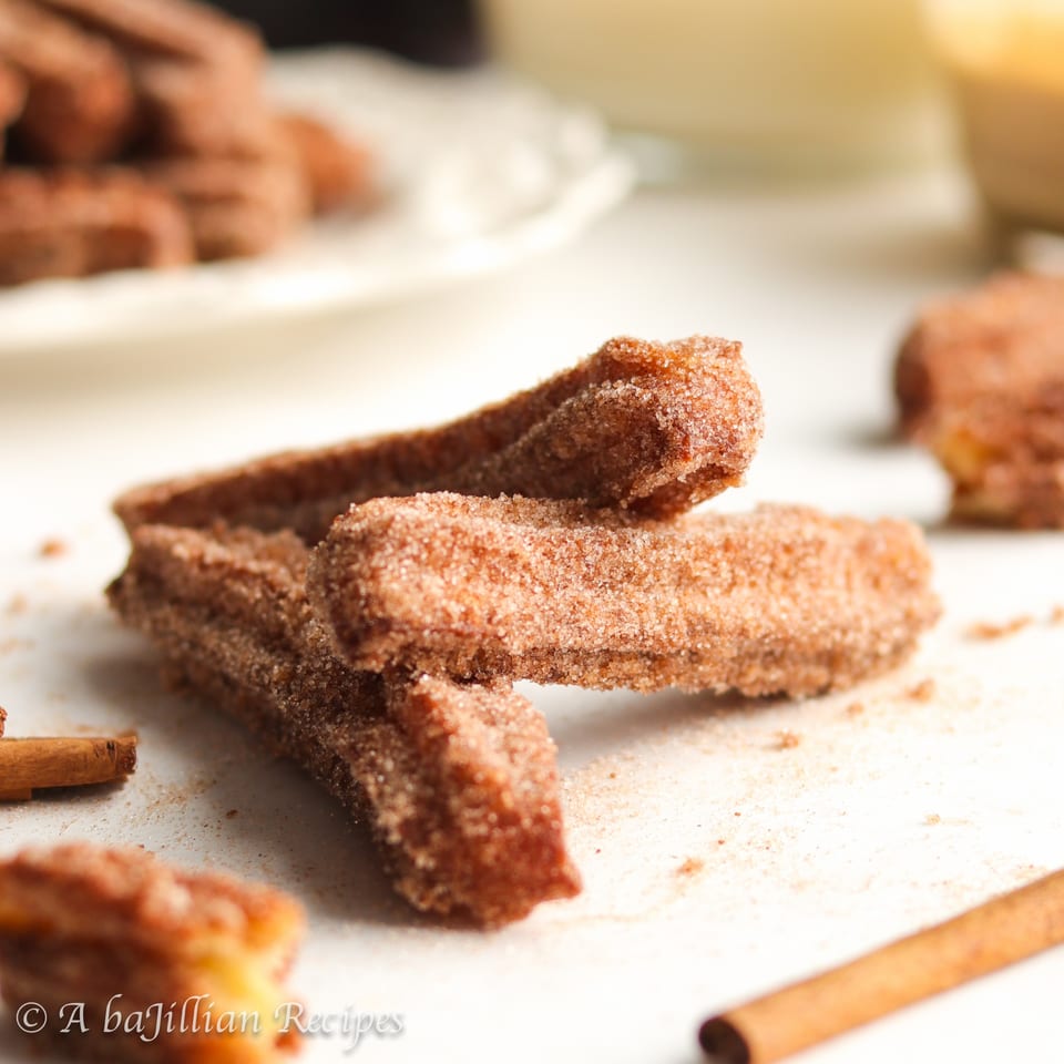 Sugar-coated cinnamon churro sticks stacked on a plate, with cinnamon sticks and blurred pastries in the background.
