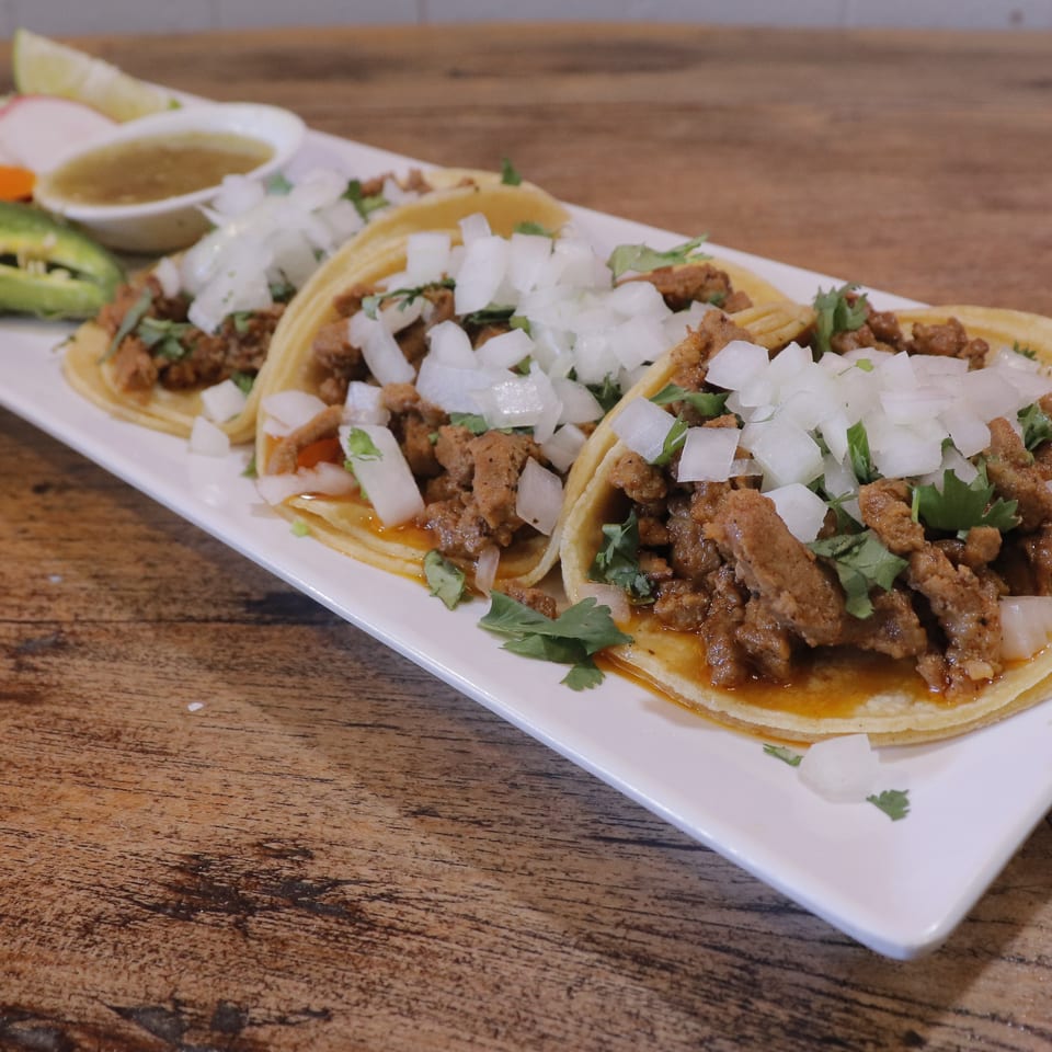 Three corn tortilla tacos topped with seasoned meat, diced onions, and cilantro, served with salsa, lime, radishes, and peppers.