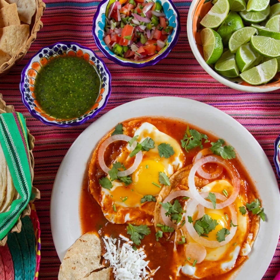 Mexican breakfast plate with eggs in red sauce, tortillas, chips, lime wedges, salsas, peppers, and colorful woven textiles.