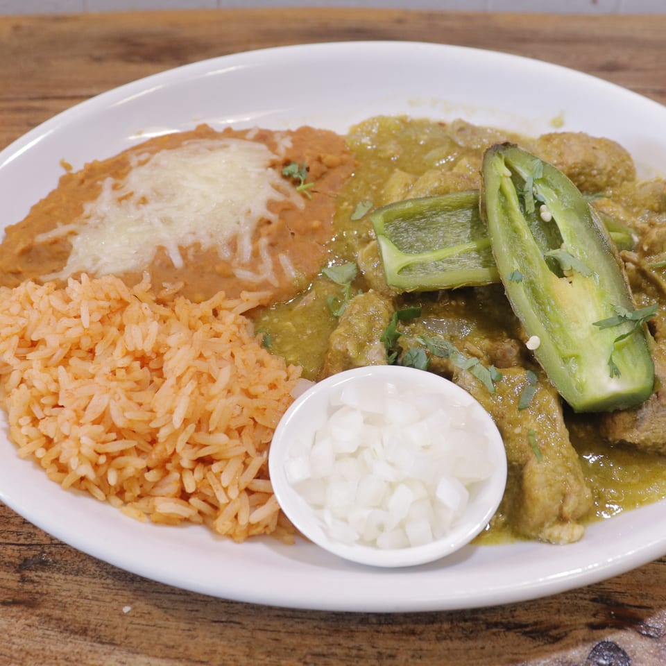Plate of Mexican-style green chile pork, orange rice, refried beans with melted cheese, sliced peppers, and diced onions.