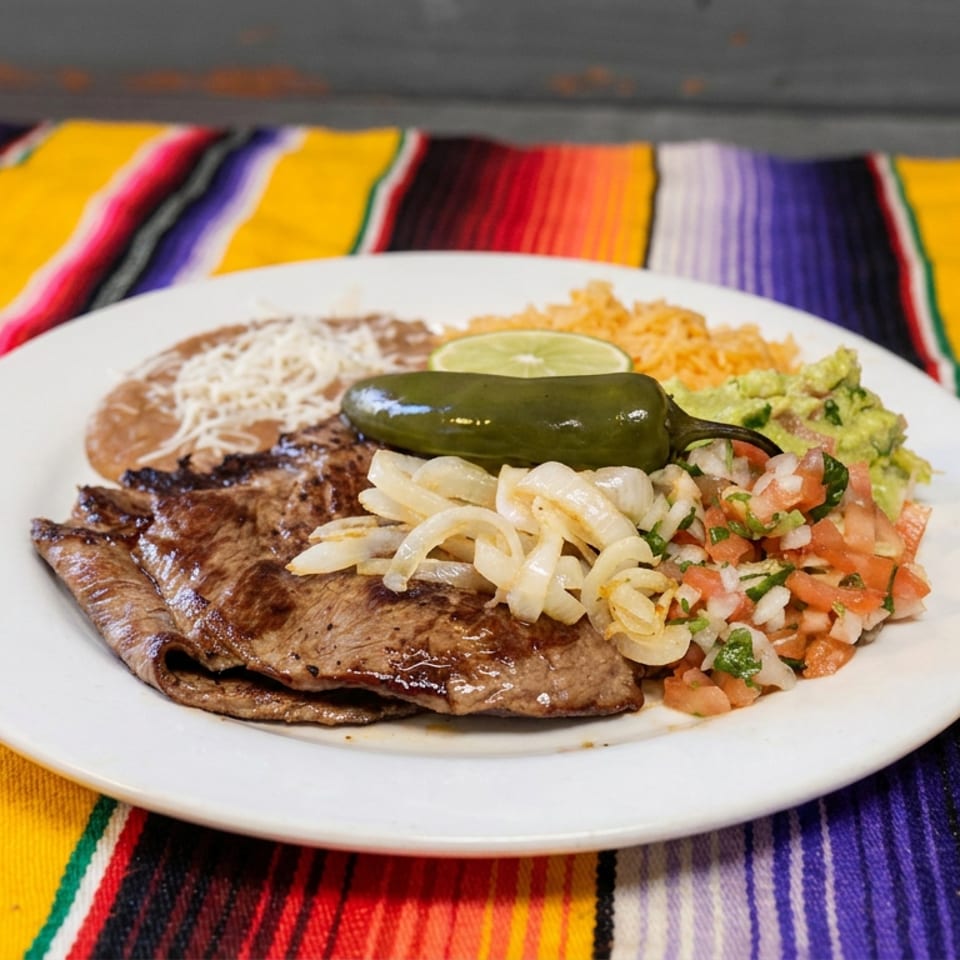 Grilled steak served with sautéed onions, refried beans, rice, guacamole, pico de gallo, and jalapeño on a colorful striped tablecloth.