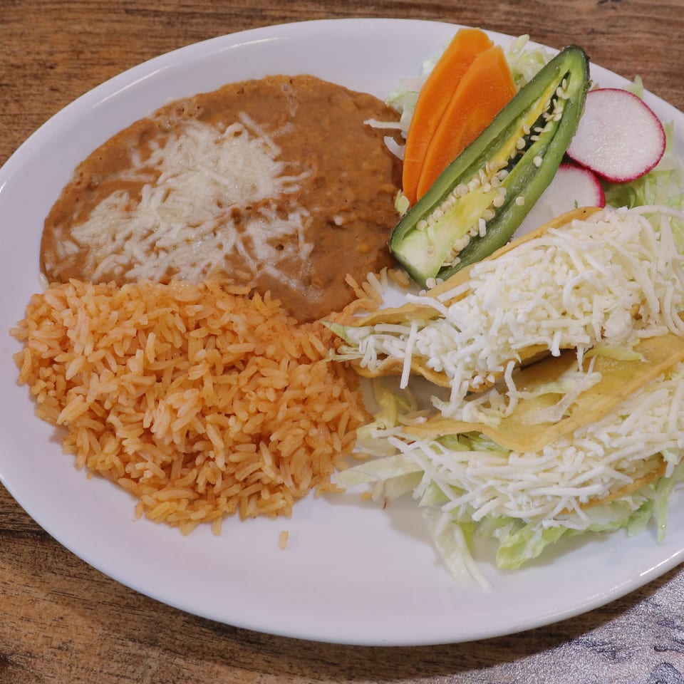 Mexican-style plate with rice, refried beans topped with cheese, shredded lettuce, crispy tacos, carrots, jalapeño, and radishes.