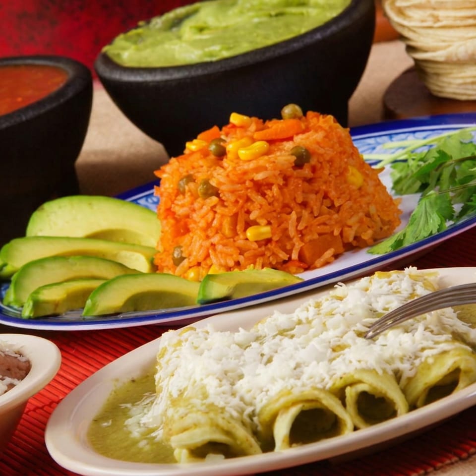 Mexican meal spread featuring green enchiladas topped with cheese, orange rice, beans, guacamole, salsa, avocado, and tortilla chips.